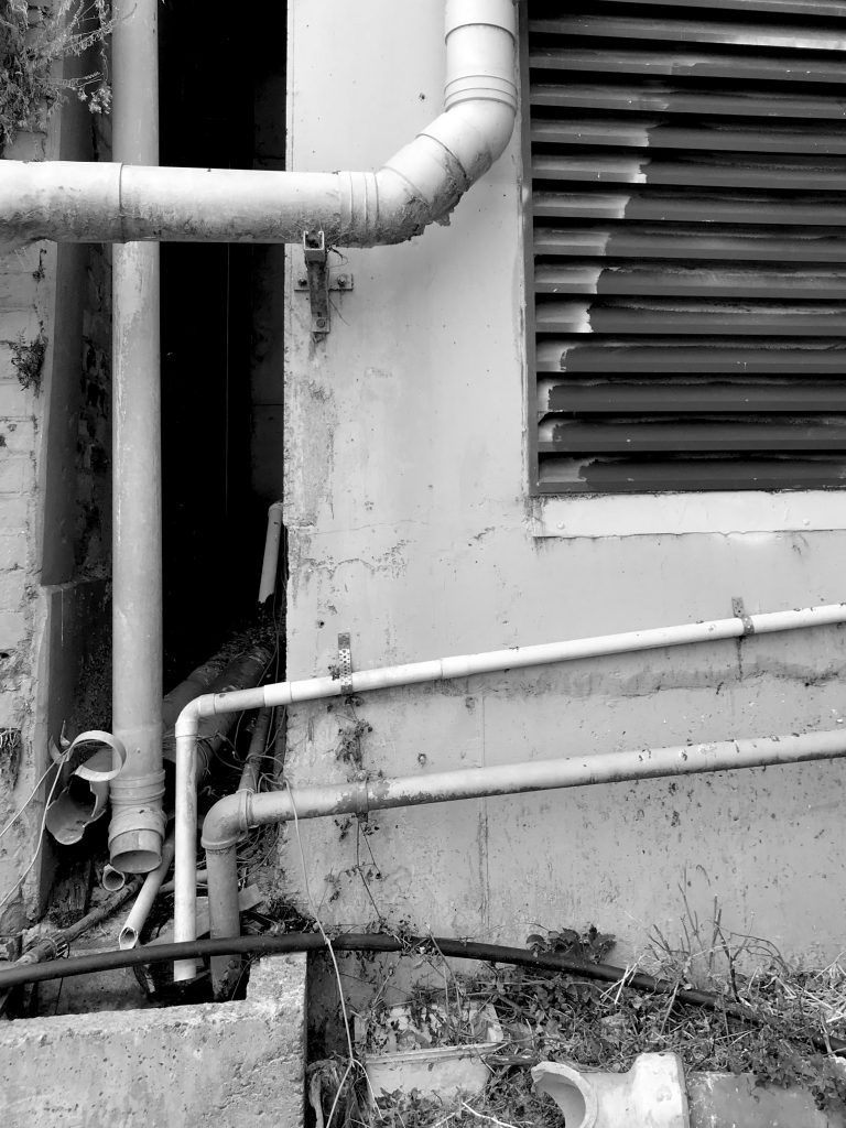 A black and white photograph of a worn wall with pipes and a vent