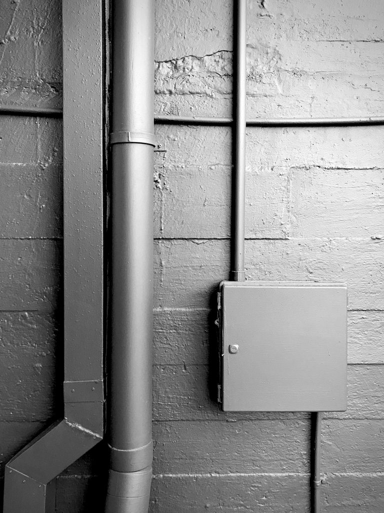 A black and white photograph of a power box fixture and drain pipes on a dark wall