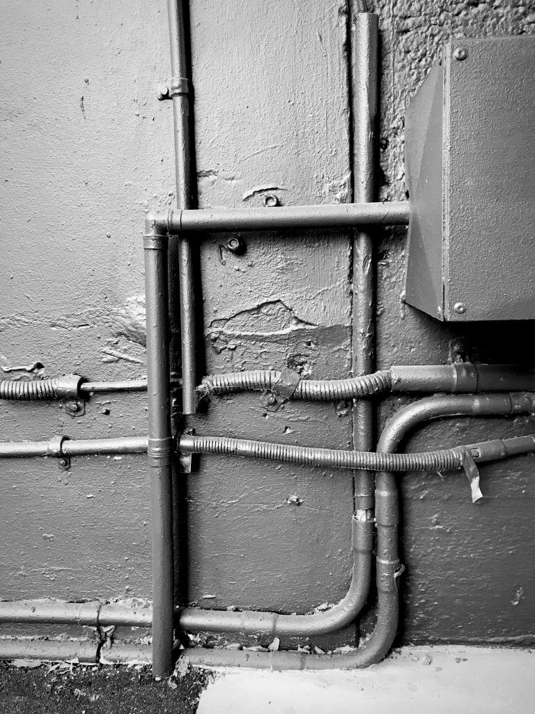 A black and white photograph of a group of pipes running across a wall, the pipes and the wall have been painted in a dark tone.