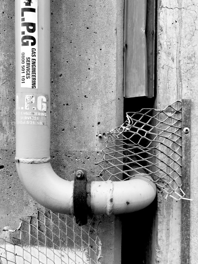 A black and white photograph of a pipe emerging from a wall with a metal grill surrounding it.