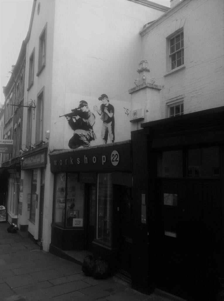 A famlous stencil by Banksy, an army sniper takes aim from above a shop, but behind him a child holds a paper bag, blown up and ready to surprise the sniper with a bang