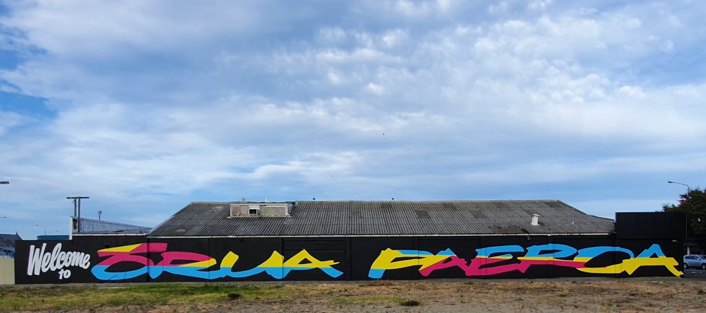 A long block wall is painted black, with the words Welcome to Orua Paeroa painted in bright colours.