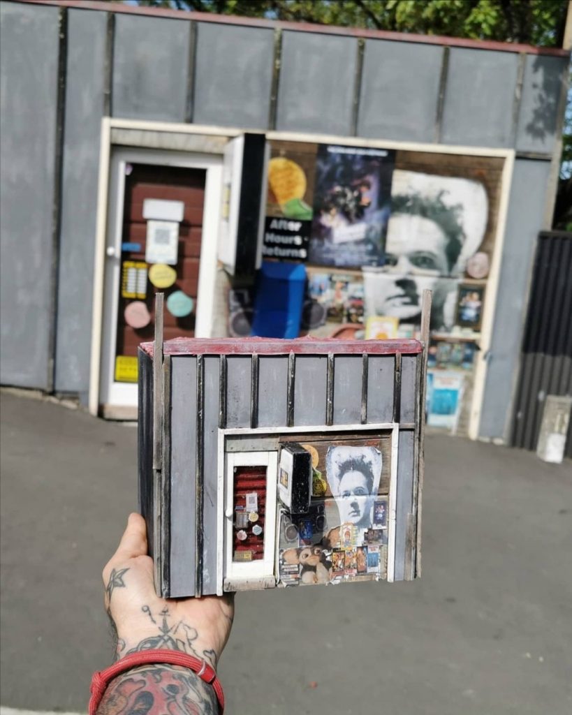 The artist holds his scratch build of the original home of Lyttelton's Dead Video in front of the real thing