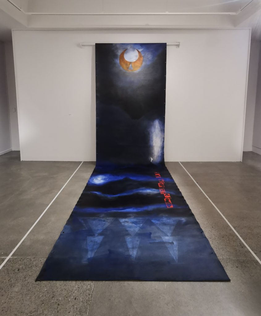 Work's piece for the TMD: An Aotearoa Graffiti Story at Lower Hutt's Dowse Art Gallery. The unfurling piece is dark blue and depicts a moon glowing at the top of the section on the wall, there are figures on the section rolled along the floor.